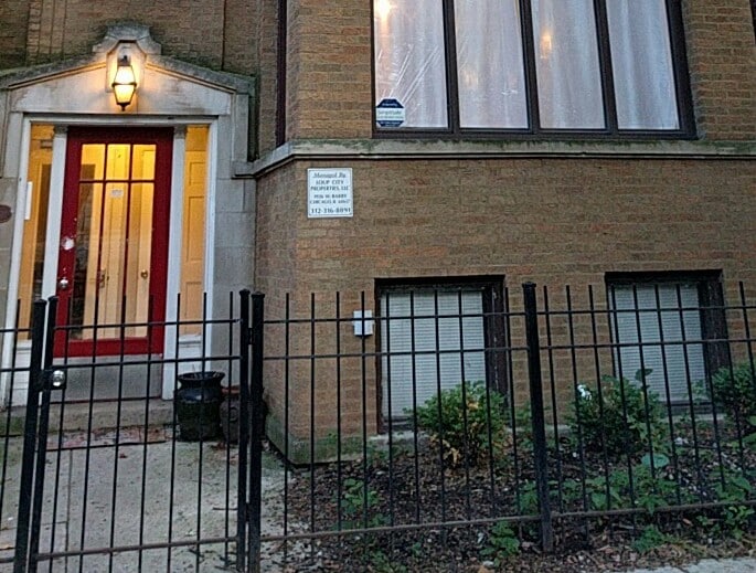 The exterior of a garden unit is visible, featuring a red door framed by large windows. A locking fence surrounds the entrance, with low shrubs and a small area of gravel alongside the pathway. The building's brick facade provides a classic urban feel.