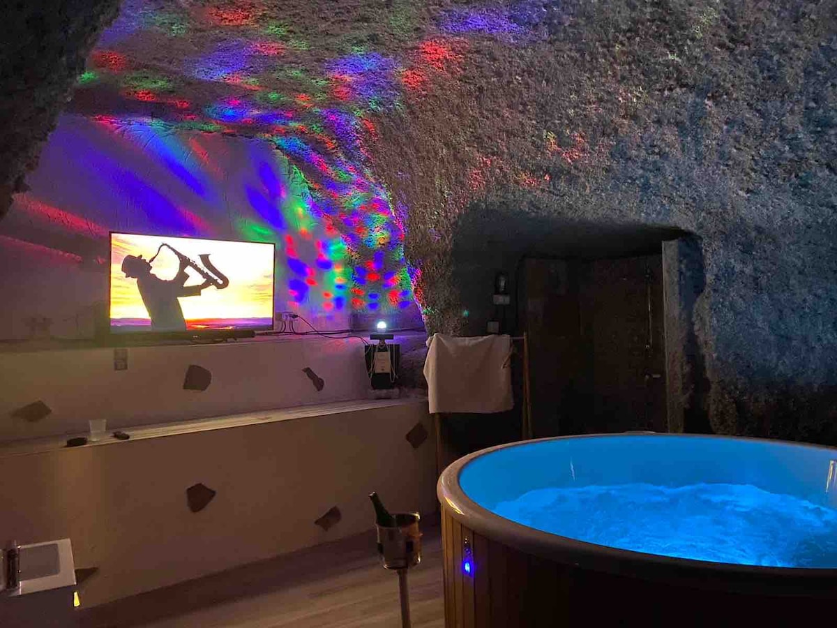 A Nordic jacuzzi bathtub is situated in a cozy cave with colorful lighting effects on the ceiling. A flat-screen television displays a relaxed scene, while wooden accents provide a natural touch to the unique space.