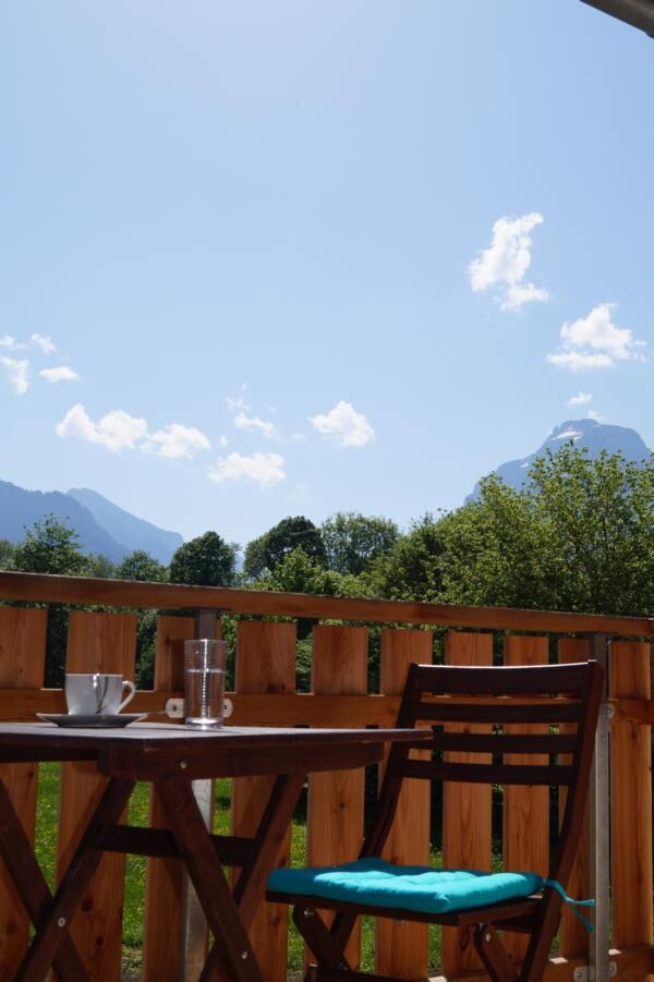 A wooden balcony table is set with a cup and glass, overlooking a scenic view of mountains and greenery. The clear sky and distant mountains can be seen in the background, creating an open atmosphere. A wooden railing frames the serene landscape.