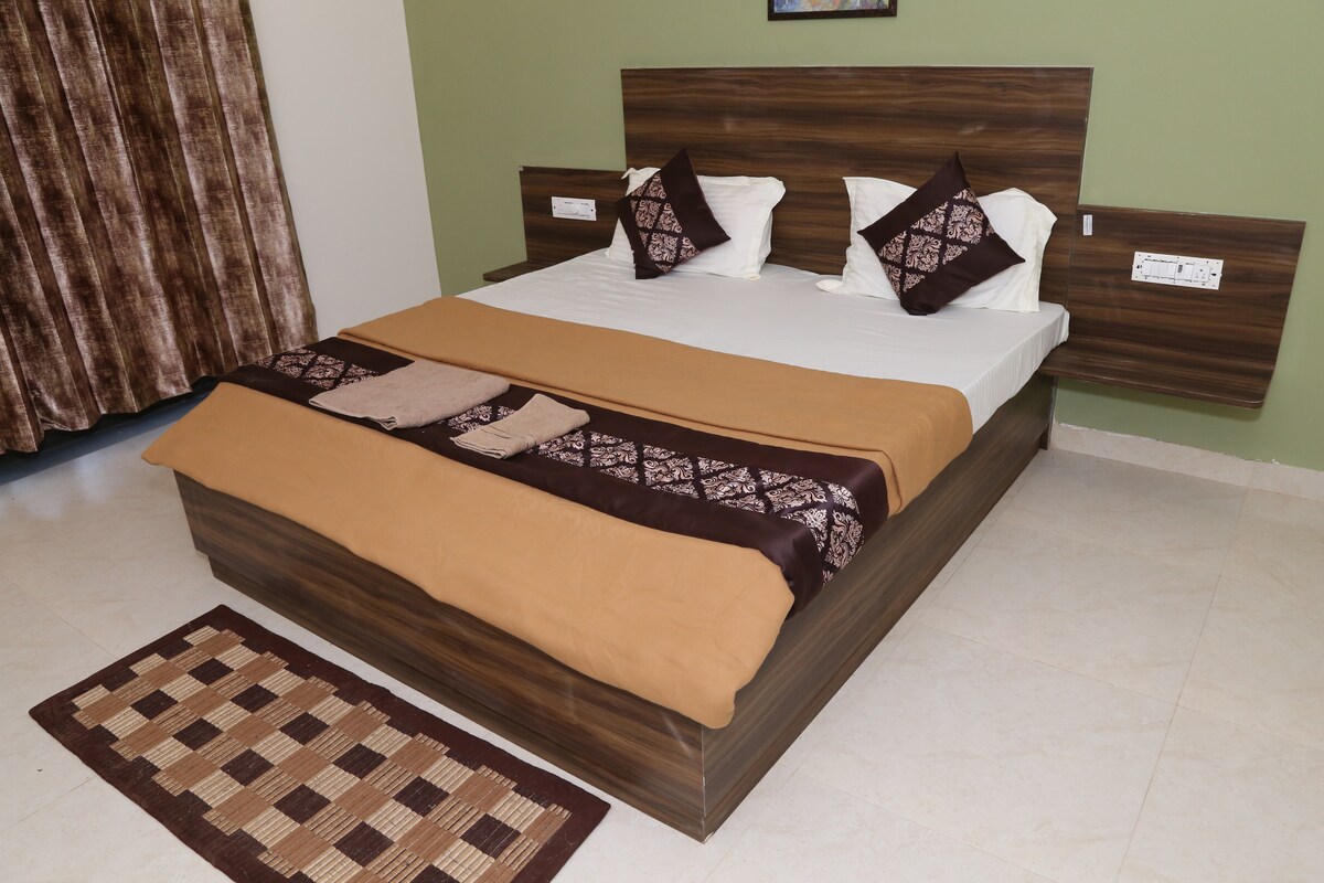 A spacious Deluxe room is highlighted by a King-sized bed, adorned with neutral-colored bedding and decorative pillows. The room features tile flooring and is complemented by a simple area rug. Natural light enters through a window, accompanied by brown curtains.