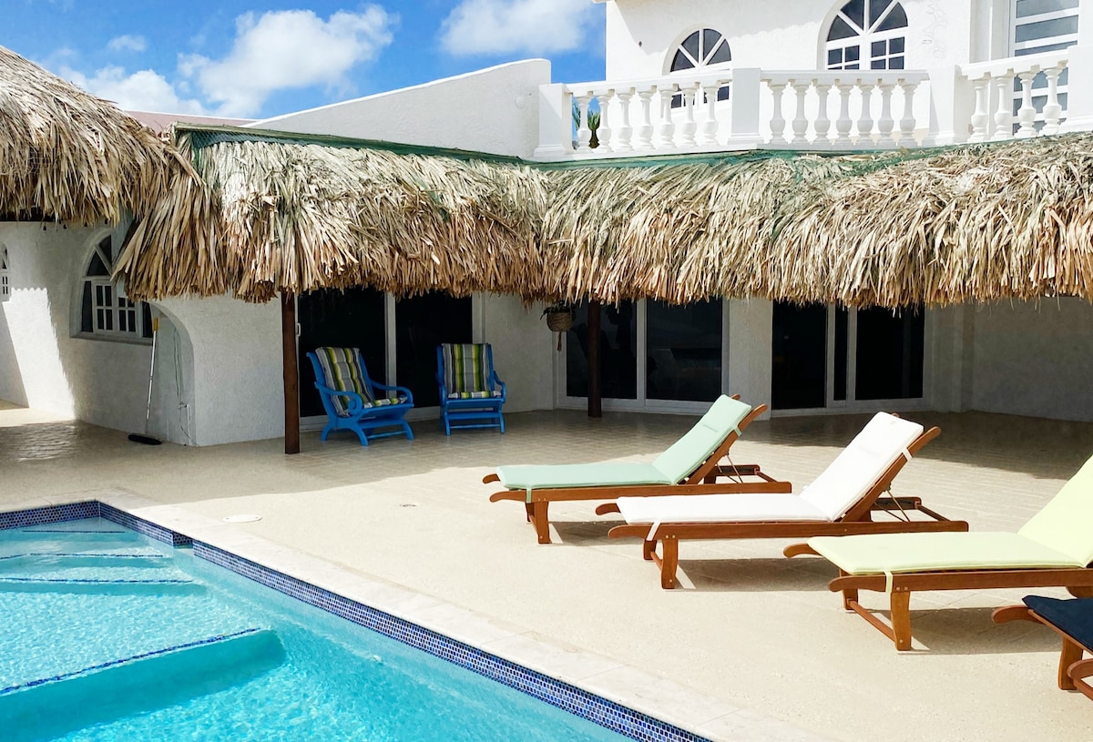 An outdoor space is shown, featuring a shimmering pool surrounded by lounge chairs in varying colors. A thatched roof offers shade, while large glass doors provide access to a well-lit interior. Decorative elements, such as tropical plants, add to the serene setting.