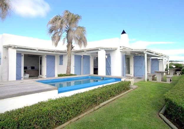 A modern cottage exterior is shown, featuring a swimming pool and a manicured lawn. Light blue doors complement the white walls, and a palm tree adds a touch of greenery beside the outdoor seating area.