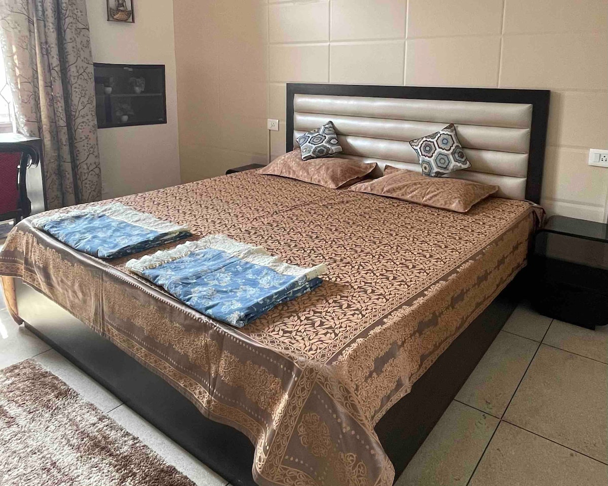 A spacious bed is dressed with a richly patterned bedspread and two decorative pillows. Each side of the bed is accompanied by a nightstand, and light enters through a window dressed with patterned curtains. A rug adds warmth to the tiled floor.