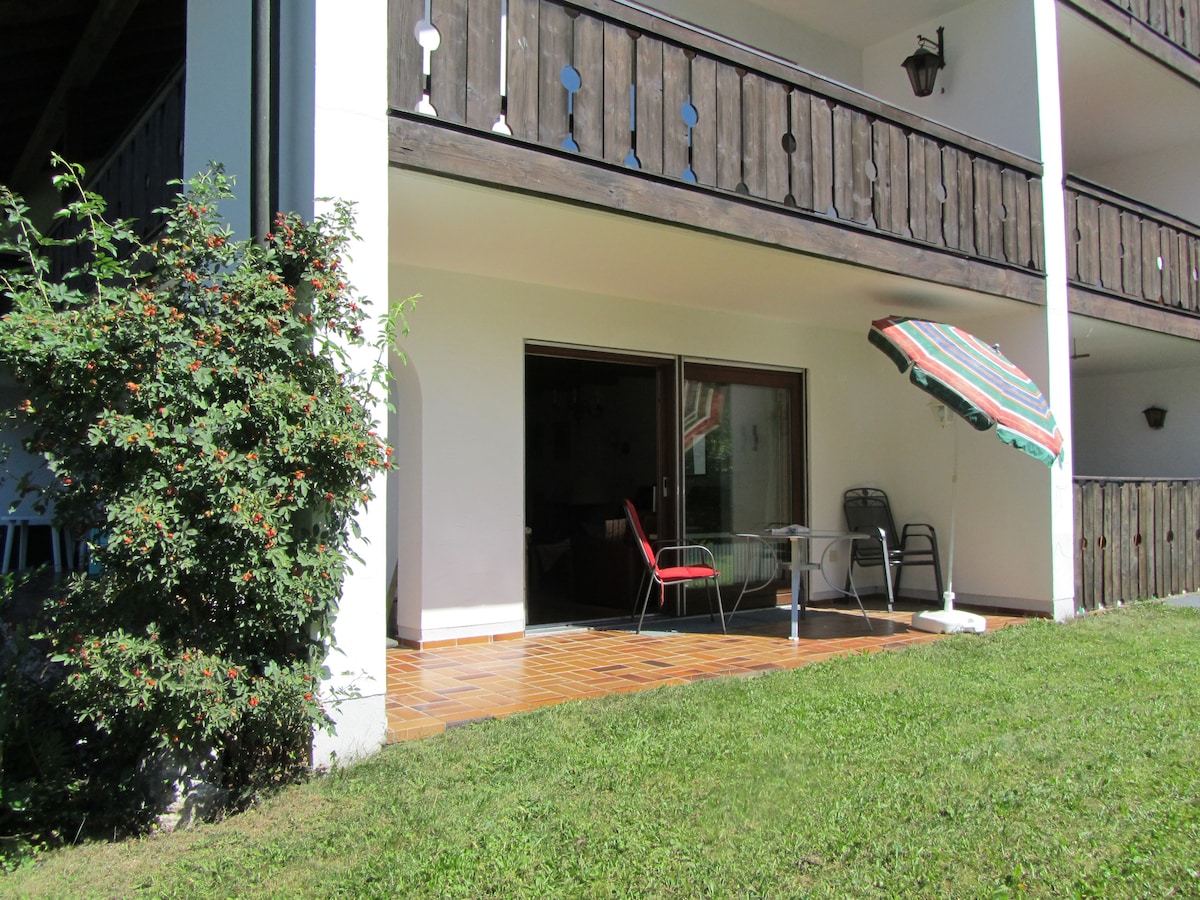 The terrace features outdoor seating with a table and chairs under a colorful awning. Lush green grass lies in front of the terrace, with a shrub partially visible nearby. Large sliding doors lead into the indoor living space, allowing natural light to fill the area.
