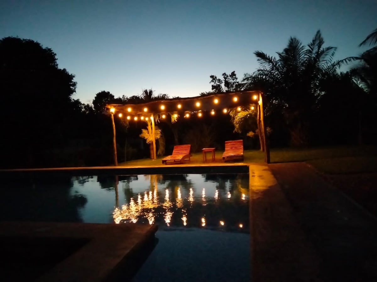 An illuminated pool area is featured, with warm string lights draping over a wooden structure. Two lounge chairs are positioned beside the pool, reflecting the shimmering water beneath the evening sky, surrounded by lush greenery and palm trees.