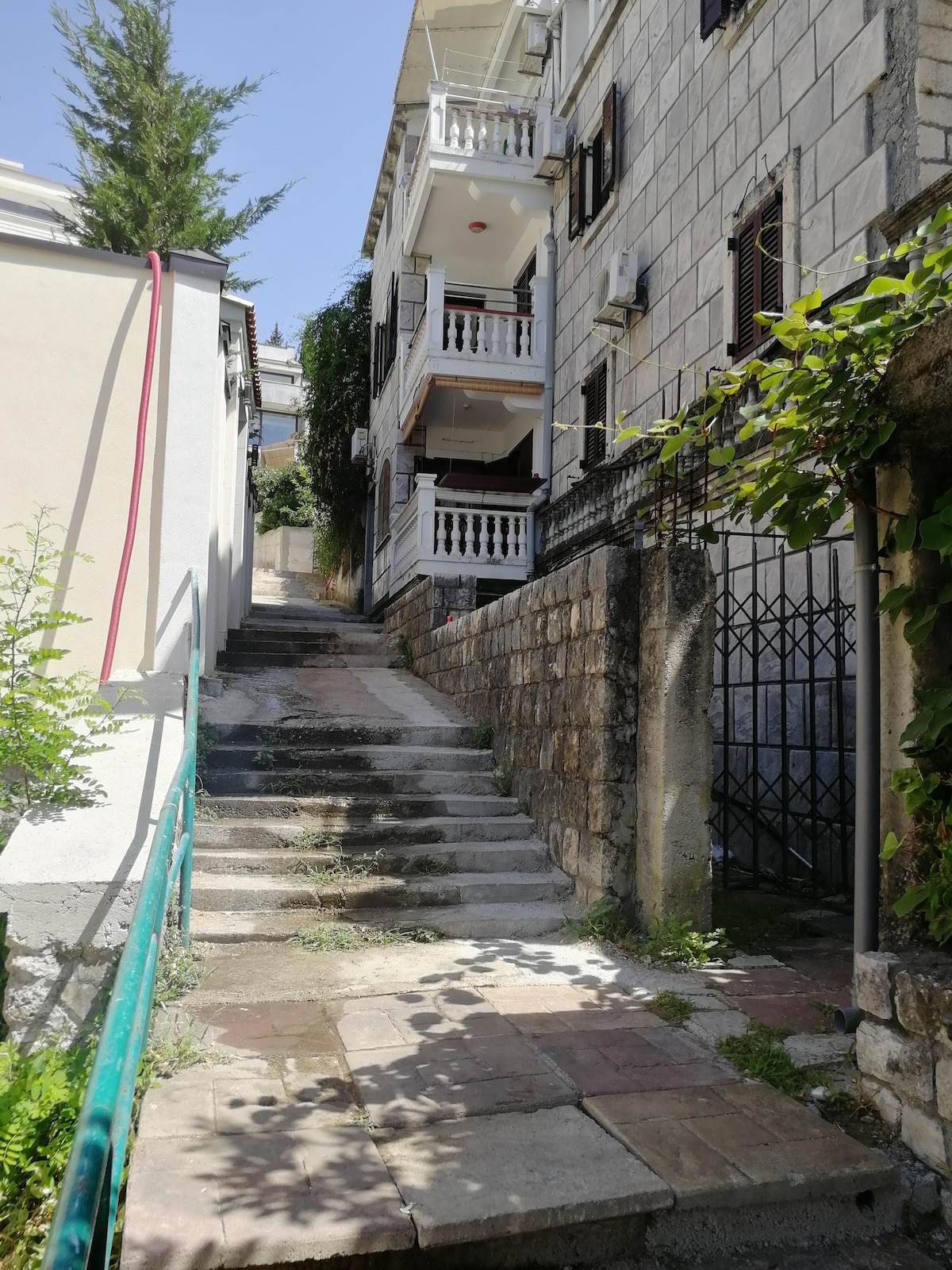 A stone pathway leads up a gently sloping incline, bordered by green foliage and a railing. Buildings with light-colored stone walls are visible in the background, showcasing balconies adorned with white railings. Sunlight casts soft shadows on the steps and surrounding surfaces.