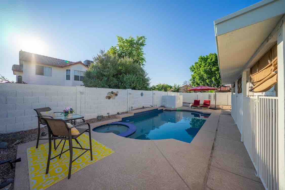 A private backyard is showcased, featuring a heated pool surrounded by a concrete patio. Red lounge chairs are positioned near the pool, complemented by a small table with decorative items. Lush greenery and a shaded area enhance the tranquil setting, with a fence providing privacy.