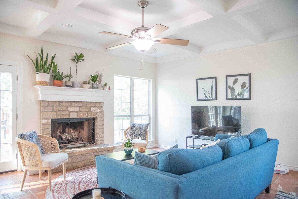 A light-filled living room features a cozy blue sofa positioned around a coffee table. A stone fireplace is framed by indoor plants, while a television rests on a sleek stand. Natural light streams through large windows, highlighting the simple decor of framed wall art.