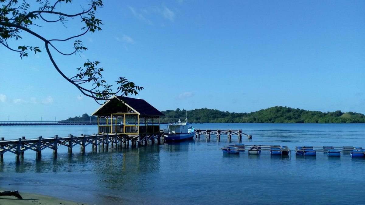 A wooden pier extends over calm waters, leading to a covered fishing shelter. A boat is moored nearby, surrounded by a few small floating structures. Lush green hills rise in the background, under a clear blue sky.