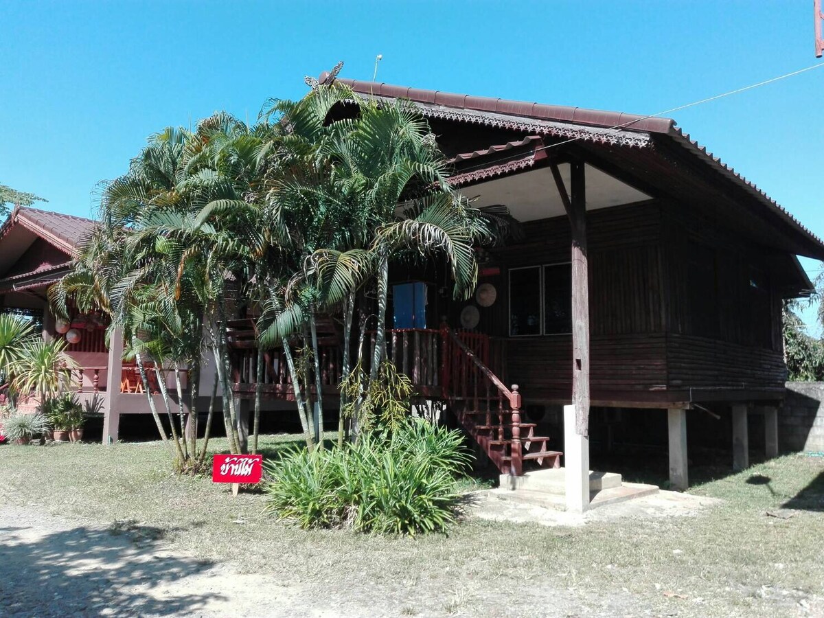 A traditional wooden house is presented with a gabled roof and a elevated porch. Lush green plants surround the exterior, adding to the natural environment. The sunlit facade features wooden railings and an inviting entrance, creating a warm welcome for guests.