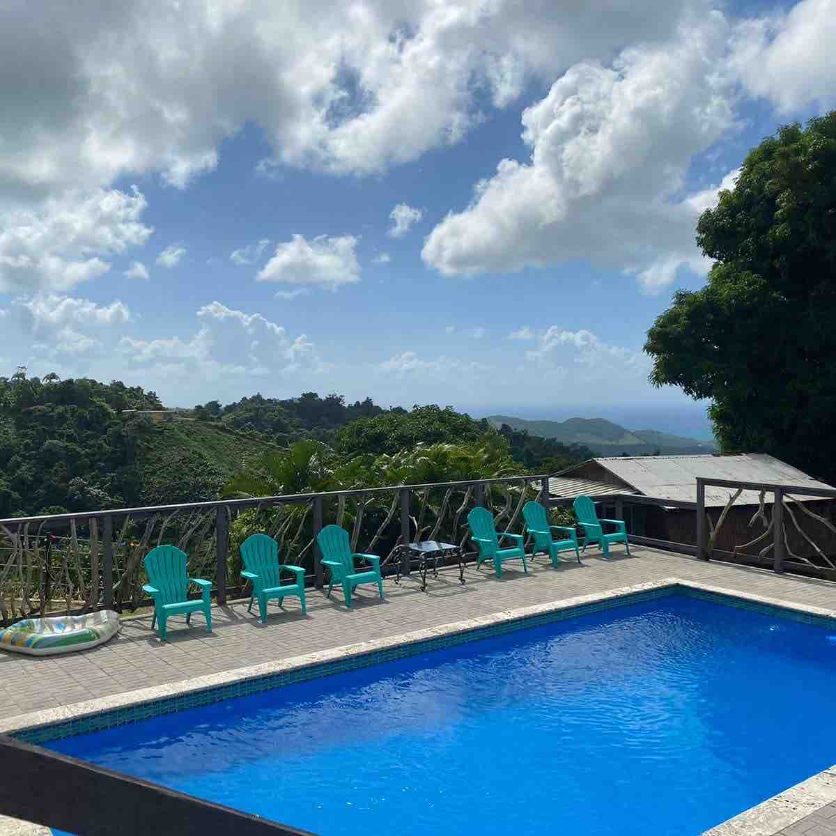 A bright outdoor pool is framed by several turquoise lounge chairs, providing a relaxing space. Lush greenery and rolling hills are visible in the background, under a partly cloudy sky that enhances the serene atmosphere.