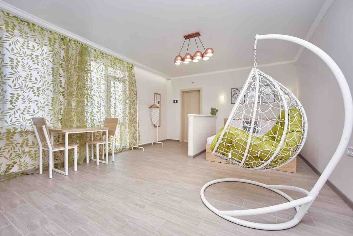A cozy living space features a unique hanging chair with a vibrant green cushion, adding a playful touch. A small dining table with two chairs is positioned nearby. Large windows are adorned with light, leafy curtains that allow natural light to fill the room.