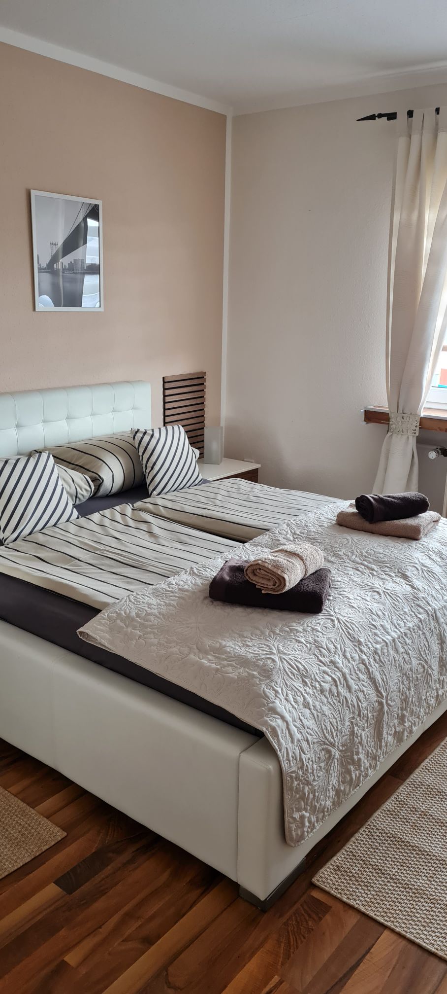 A bright bedroom features a spacious bed with a light-colored quilt and striped pillows. Soft towels are neatly placed on the bed and beside a wooden nightstand. Natural light filters through sheer curtains, illuminating the warm wooden floor and enhancing the calming atmosphere.