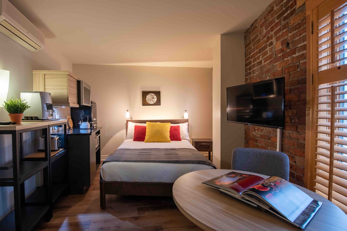 A cozy studio room features exposed brick walls, enhancing its rustic charm. A double bed with colorful pillows is positioned alongside a small round table. A kitchenette with basic amenities is visible, along with a smart TV mounted on the wall, providing a functional living space.