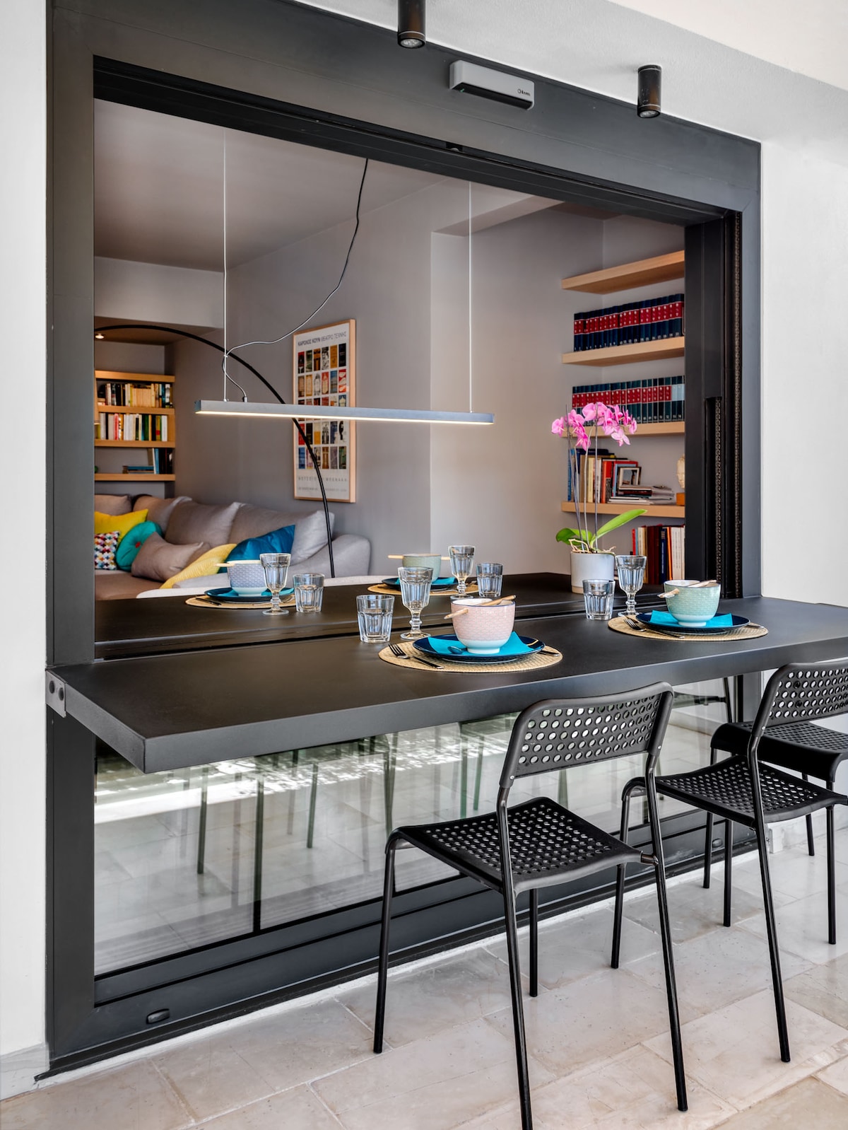A modern dining area presents a sleek black table with stylish utensils arranged for four. Transparent glass surrounds the space, allowing light to enhance the view of a cozy living room in the background, which features colorful cushions and shelves filled with books.