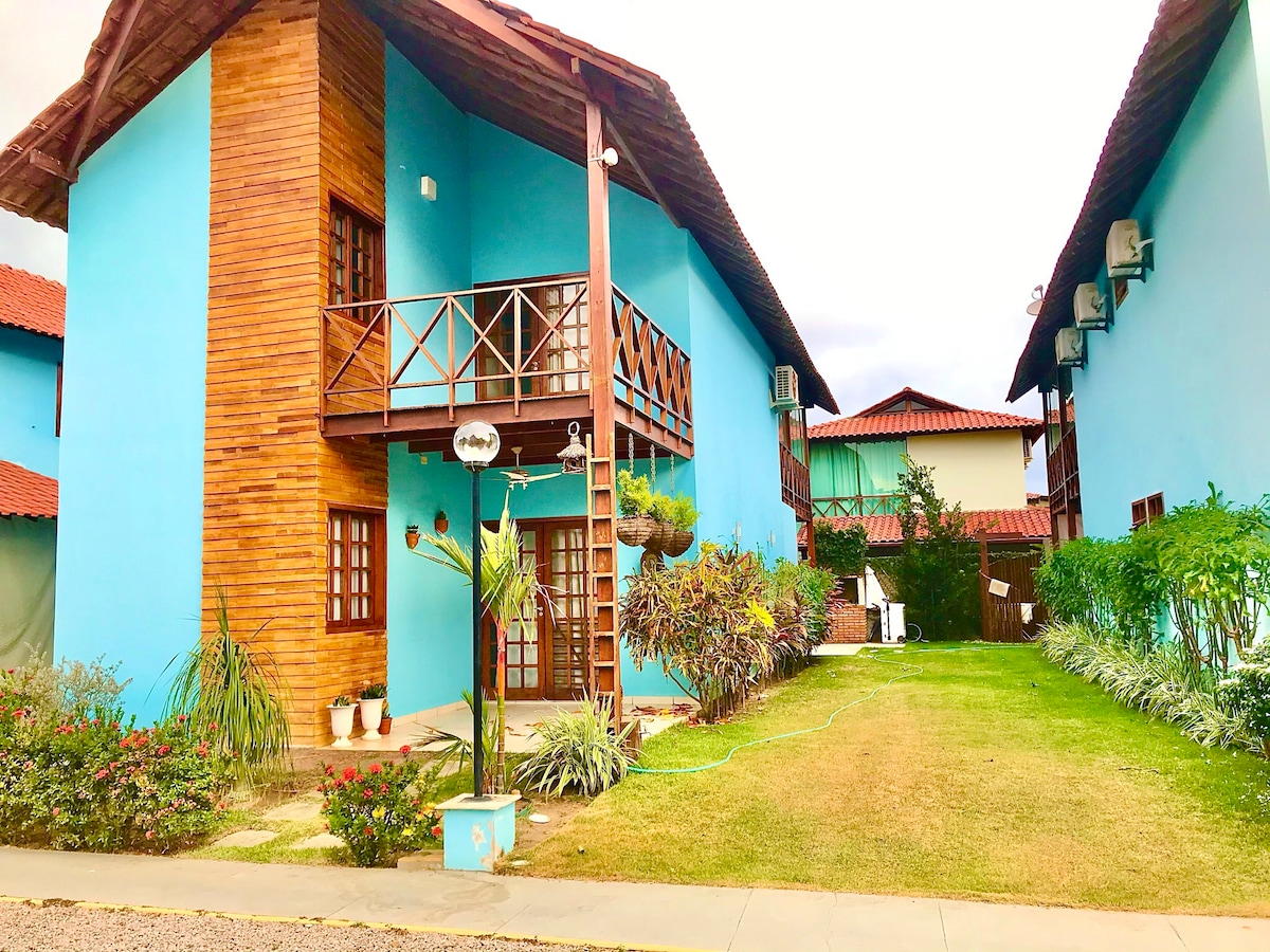 A vibrant blue exterior characterizes the two-story building, featuring wooden balcony railings and air conditioning units. Lush greenery, including potted plants and shrubs, enhances the landscaped area. A well-maintained lawn leads towards an inviting entrance, framed by a ladder and additional outdoor features.