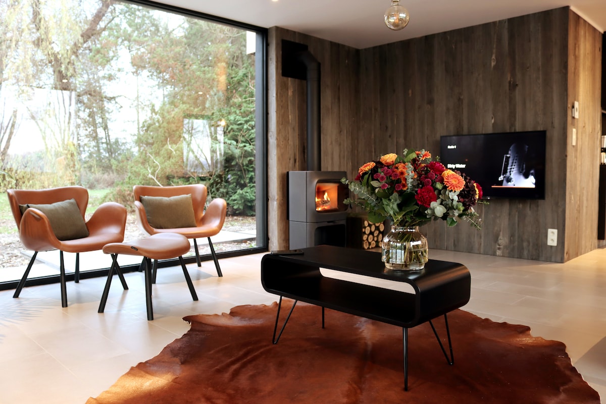 A cozy living area is highlighted by large windows that provide views of the surrounding greenery. Two stylish chairs are placed beside a dark coffee table on a textured rug. A modern fireplace is visible, accompanied by a beautifully arranged vase of colorful flowers.