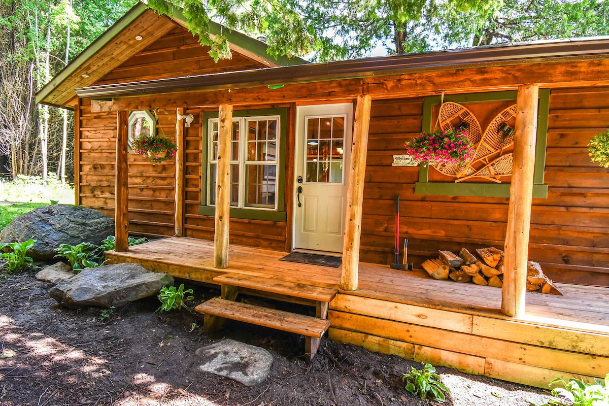 A charming cabin exterior features natural wood siding and a welcoming porch. The entrance is framed by colorful flower baskets, and a stack of firewood is neatly arranged beside the door. Surrounding greenery adds to the serene atmosphere.