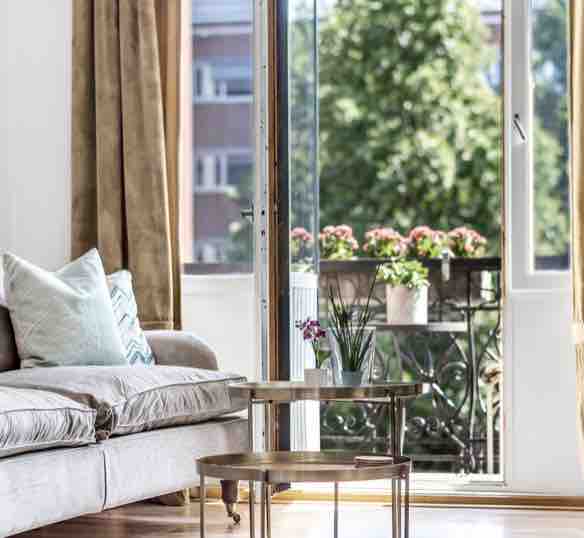 The living area features a comfortable sofa adorned with decorative pillows. A small glass table sits in front of the sofa, and natural light pours in through an open door, which leads to a balcony. Potted plants are visible outside, adding a touch of greenery.