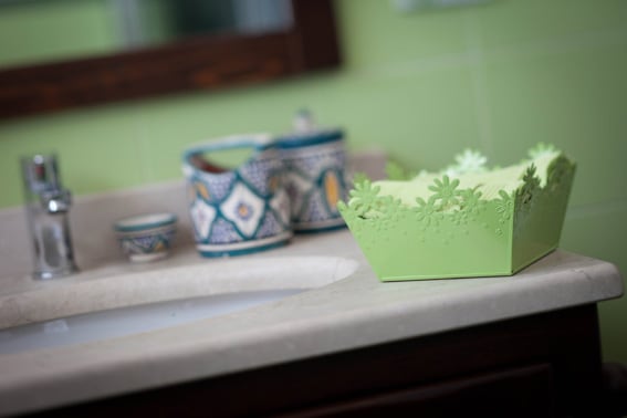 A bathroom counter is visible, featuring a light green decorative storage box with floral cutouts, along with a colorful ceramic pitcher and a small bowl. A white sink is partially seen, set against green tiled walls.