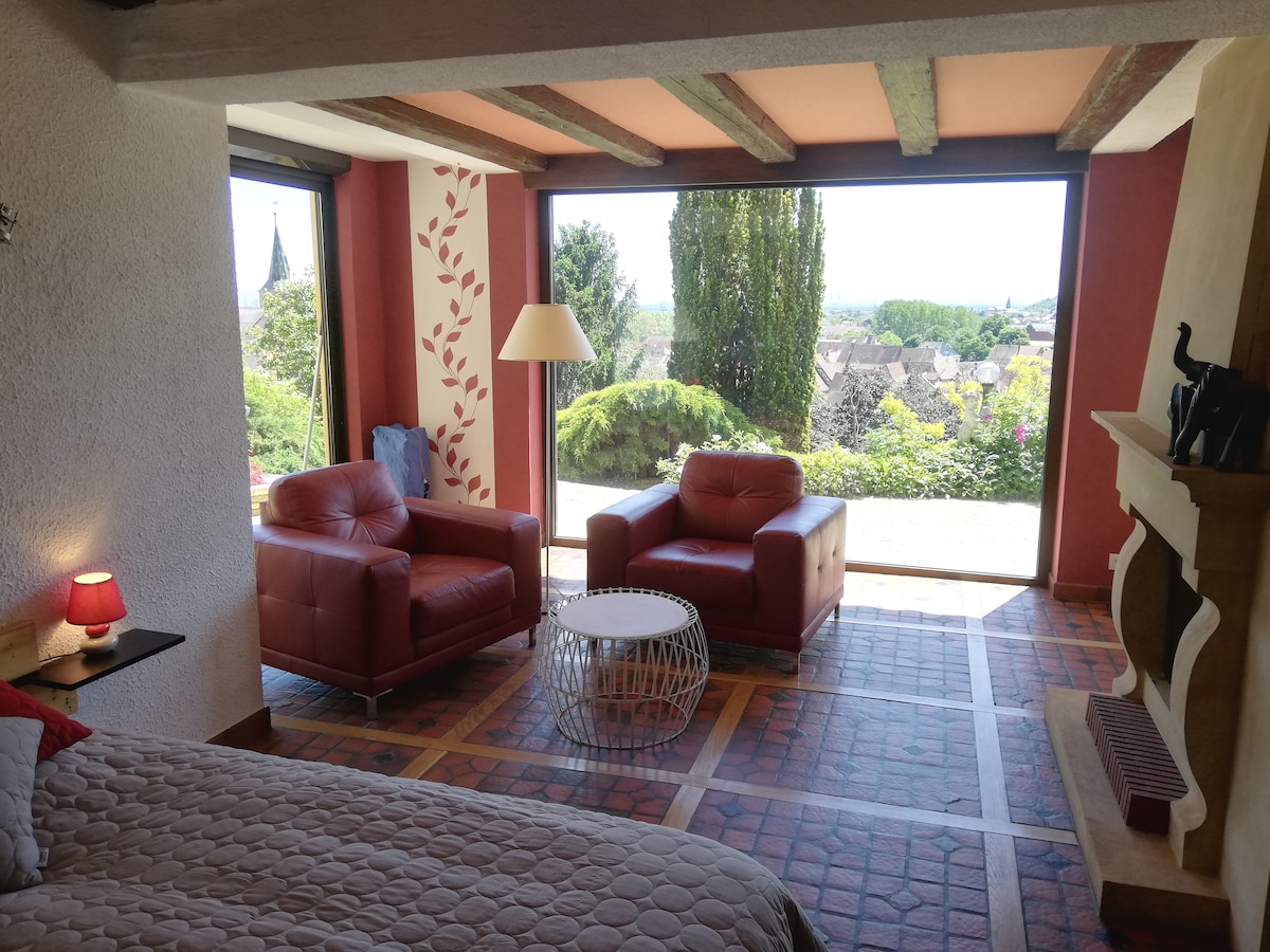 A bright living area displays two red leather chairs facing a sleek white coffee table. Large glass doors provide a view of the surrounding greenery and landscape. A soft bed is positioned nearby, and light illuminates the room through the openings.