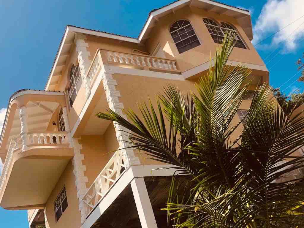 The multi-level building showcases a light-colored exterior with decorative elements around the windows. Balconies are present on upper levels, offering elevated views. Lush greenery, including palm fronds, surrounds the base of the structure, contributing to the natural setting.
