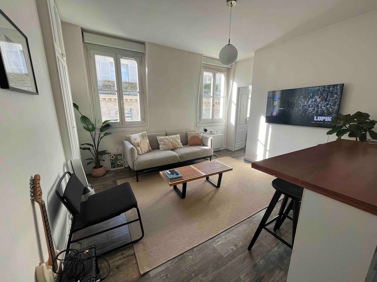 A bright living area features large windows allowing natural light to fill the space. A comfortable sofa and a low coffee table are centered on a light rug. A small dining counter with stools is visible, along with a decorative indoor plant and guitar on the floor.