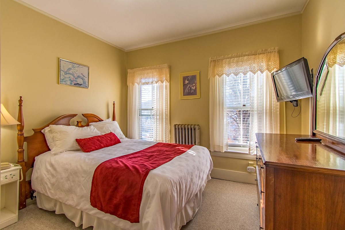 A cozy bedroom features a spacious bed dressed in white linens with a burgundy accent blanket. Natural light filters through two large windows, adorned with lace curtains. A wooden dresser and a mirror complement the warm yellow walls, while a bedside lamp adds to the inviting atmosphere.