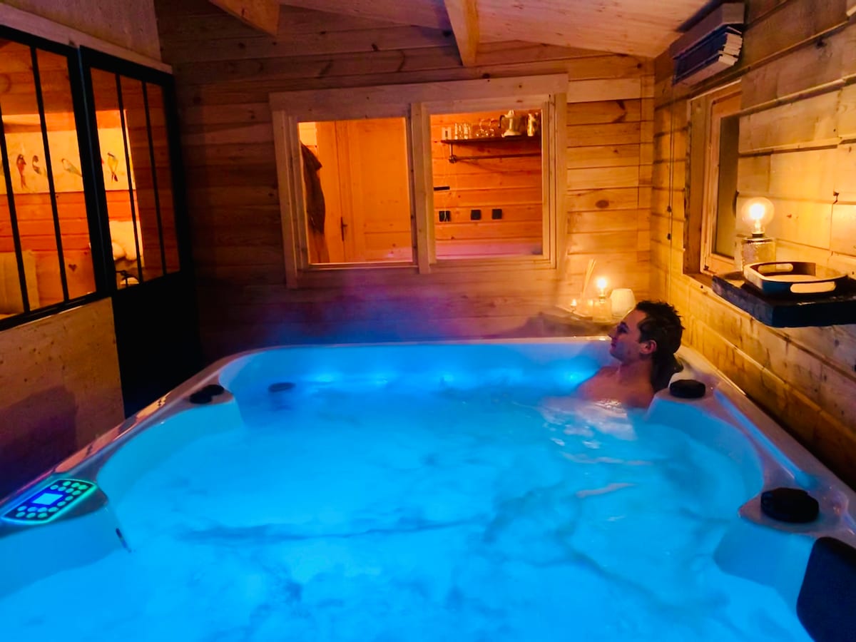 A warm, inviting space features a spacious jacuzzi illuminated in soft blue light, surrounded by wooden walls and minimalistic decor. Ambient candles provide soft lighting, enhancing the cozy atmosphere, while windows offer glimpses of the serene outdoor setting.