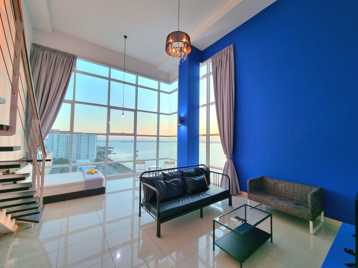 A bright living space features a stunning view of the ocean through large floor-to-ceiling windows. Comfortable seating arrangements, including a black sofa and a gray lounge chair, are complemented by striking blue walls, while soft lighting is provided by a unique chandelier.