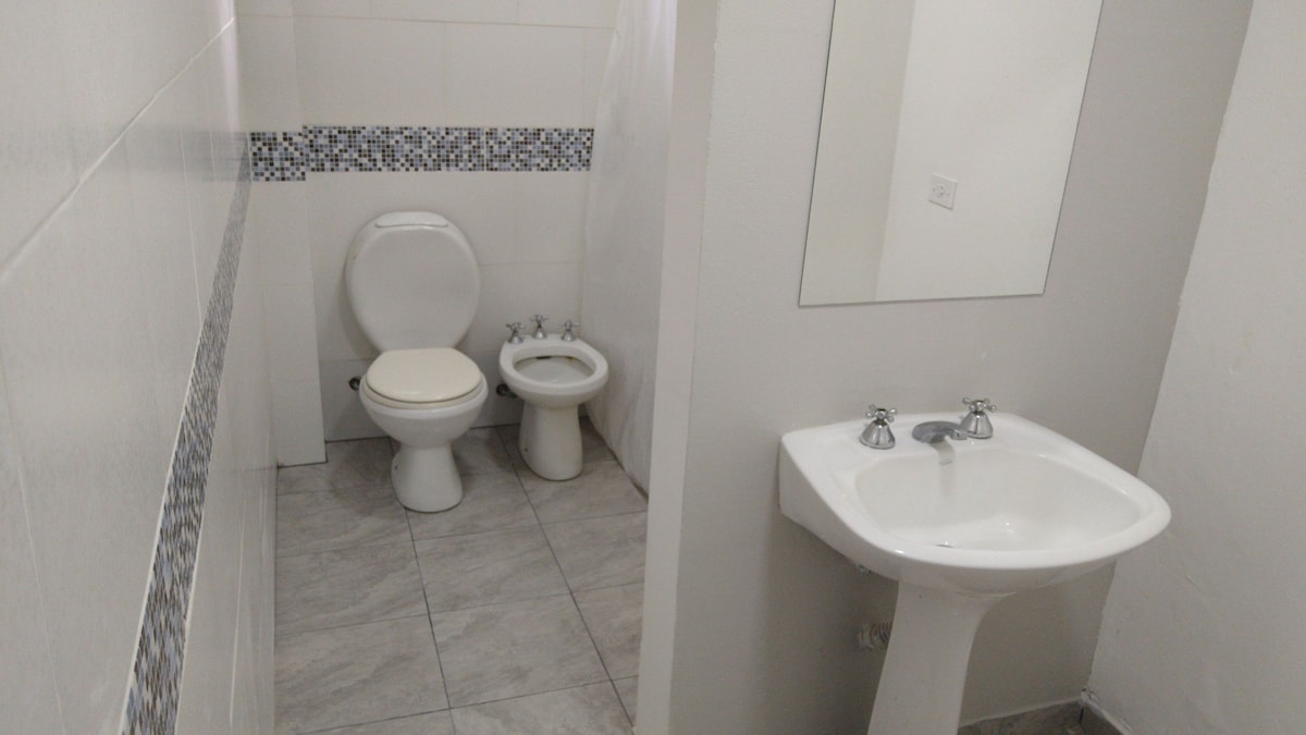 A clean bathroom space is visible, featuring white tiled walls and a mosaic accents along the middle. A toilet and a bidet are positioned beside a compact sink with chrome faucets. The floor is finished with light gray tile, enhancing the area’s brightness.