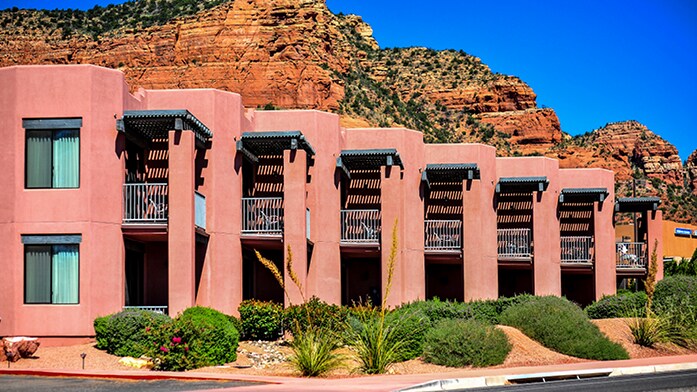 A series of distinct, rounded structures are presented in a warm terracotta color, featuring expansive windows and private balconies. Lush landscaping with native plants frames the buildings, set against a backdrop of towering red rock formations under a clear blue sky.