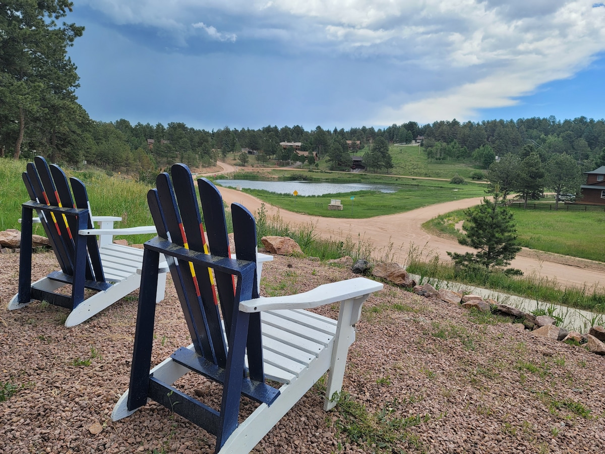 Two colorful Adirondack chairs are positioned on a gravel path overlooking a serene lake surrounded by rolling green hills and trees. The sky features varying shades of blue and gray, hinting at a tranquil atmosphere that invites relaxation.