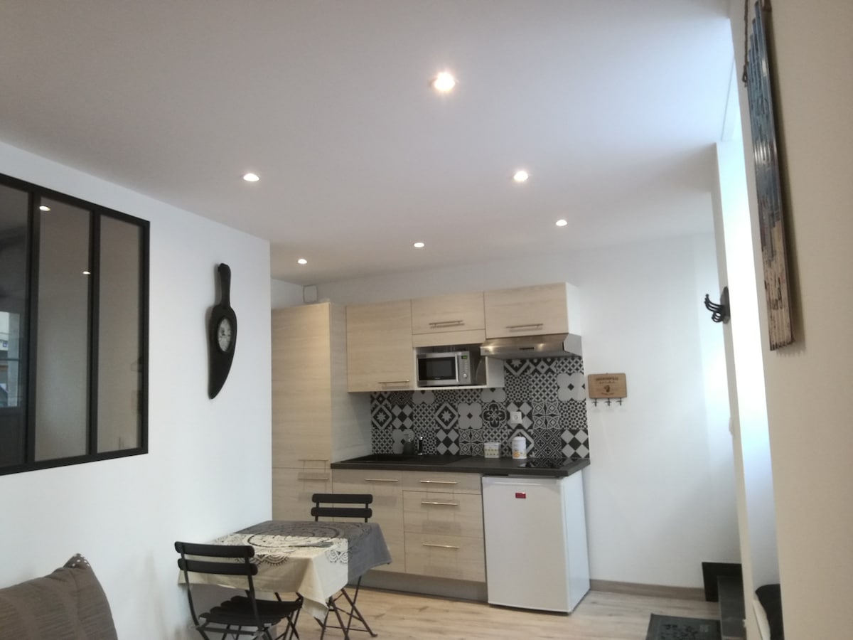 A modern studio kitchen is showcased with sleek cabinetry and a contrasting black countertop. A small dining set with two chairs is visible. Stylish tiles add visual interest to the wall. Soft lighting from recessed fixtures enhances the spacious feel of the area.