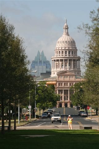 A tree-lined street leads towards the Texas State Capitol building, characterized by its iconic dome and historic architecture. The skyline of modern buildings forms a backdrop, showcasing a blend of historic and contemporary structures in an inviting urban landscape.
