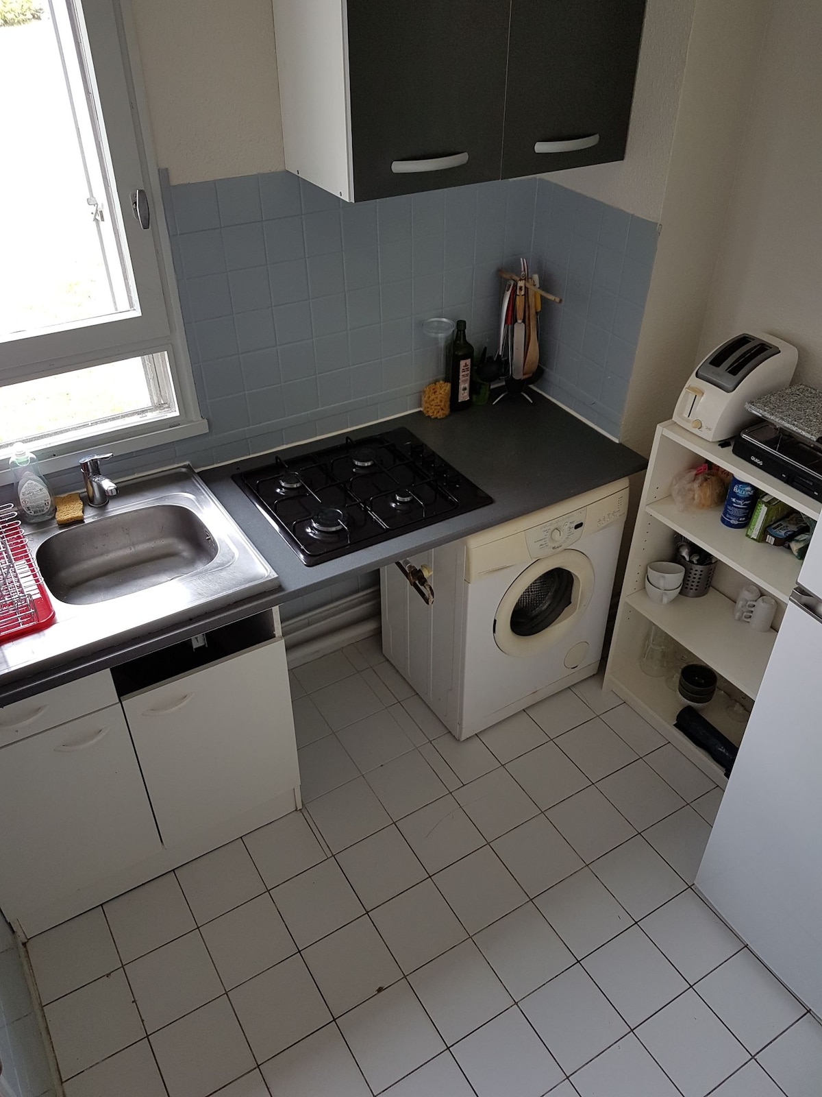 A compact kitchen area features a white washing machine next to a stainless steel sink and a four-burner gas stove. Light grey tiles cover the floor, while white cabinetry provides storage. A window allows natural light to illuminate the space.