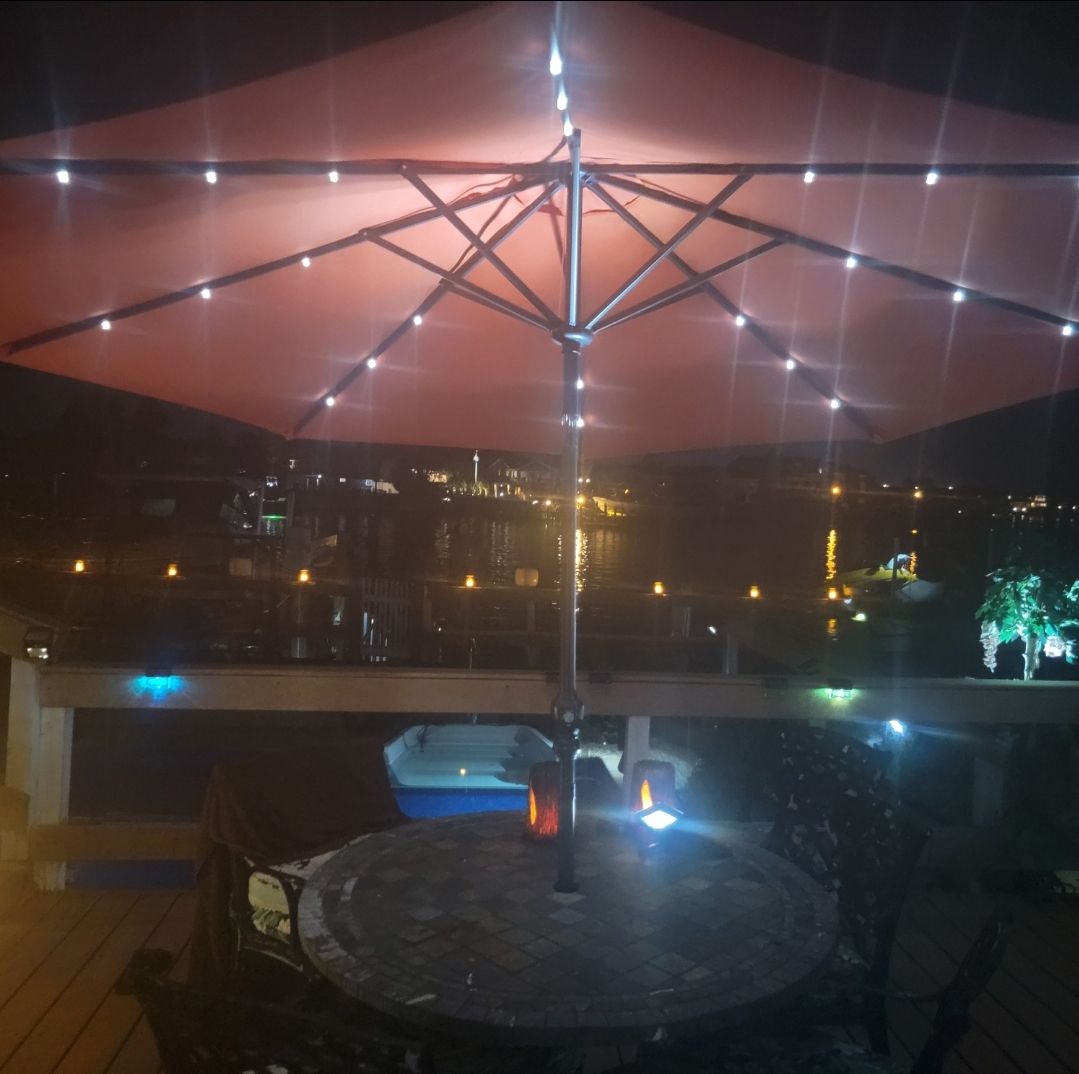 A spacious outdoor seating area is highlighted by a large, illuminated umbrella. A stone table is surrounded by four chairs, providing a cozy ambiance. In the background, the water reflects city lights, contributing to a serene evening setting.