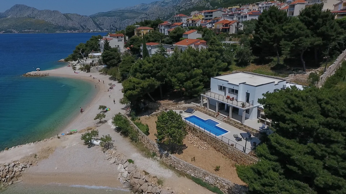Aerial view of a newly renovated house situated on a sandy beach, featuring a private pool surrounded by landscaping. The shoreline is framed by gently swaying trees, and bordered by a picturesque rocky coastline. Nearby, a beach area is visible with people enjoying the seaside.