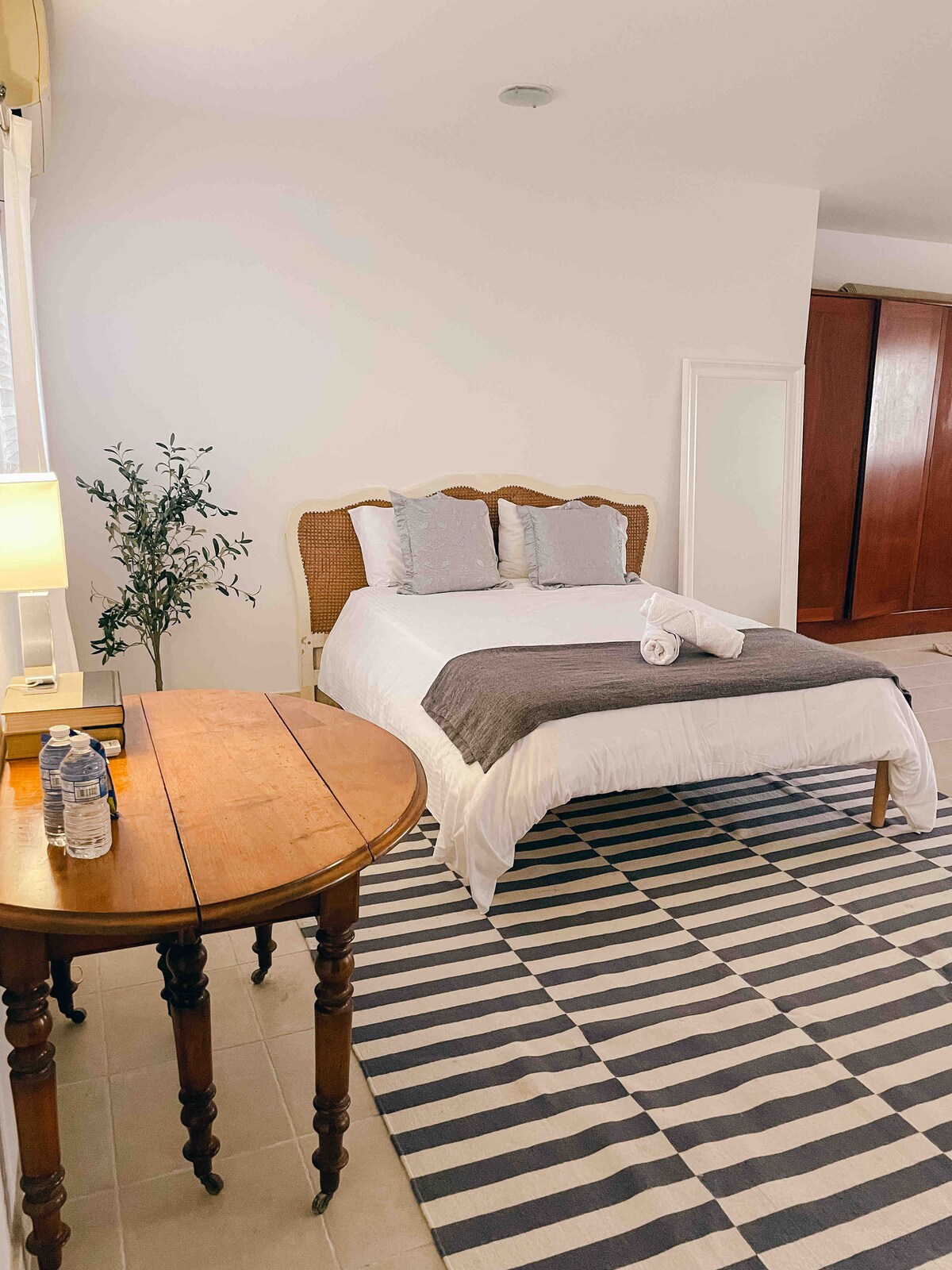 A spacious bedroom features a comfortable bed dressed in neutral linens, flanked by two pillows. A small wooden table is positioned beside the bed, with a bottle of water and a lamp. The striped rug adds a graphic element to the tiled floor.