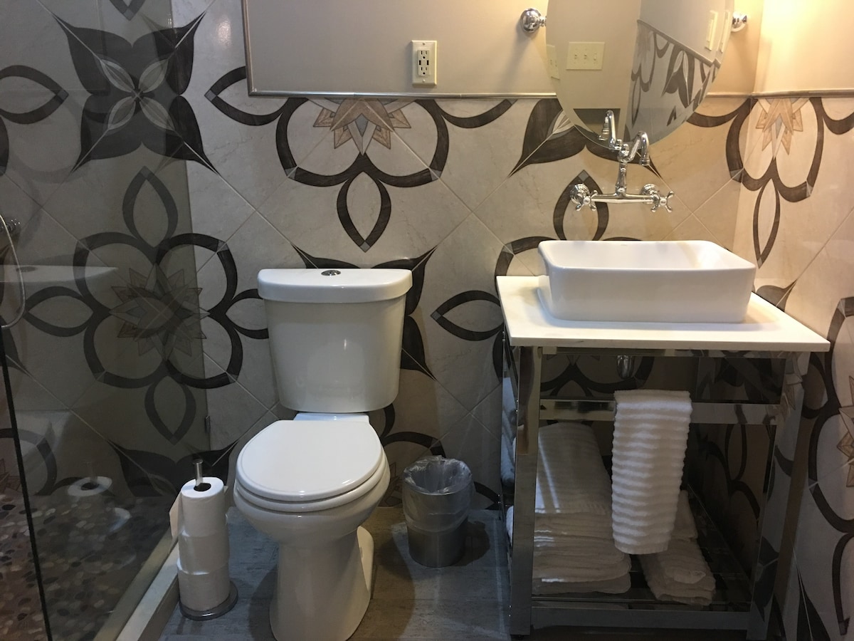 A modern bathroom features a spacious shower with glass doors and decorative tile work. A toilet is positioned beside a stylish sink, which is mounted on a metal cart. Fresh towels are neatly arranged on the cart, and a waste bin is placed nearby.