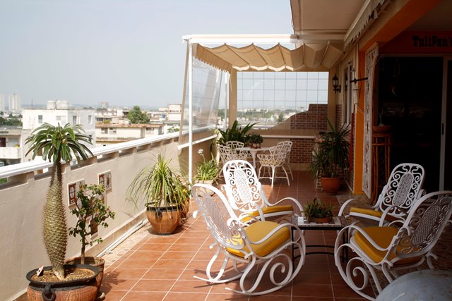 A spacious terrace is adorned with several white metal chairs featuring yellow cushions, arranged around a central table. Potted plants are placed throughout, adding greenery to the area. A protective awning covers part of the terrace, while city views are visible in the distance.