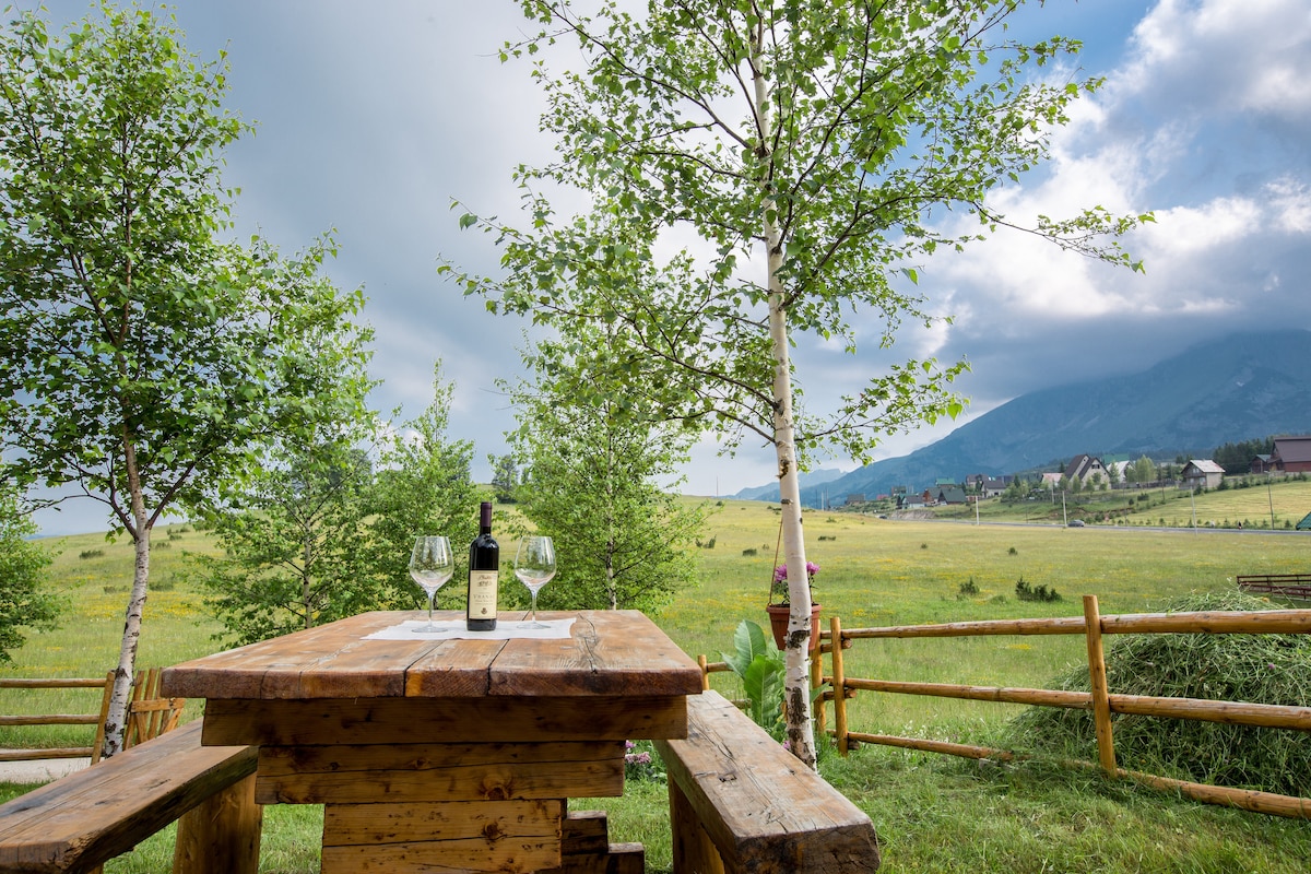 A wooden picnic table is set outside, featuring a bottle of wine and two glasses. Surrounding greenery includes trees and an expansive view of rolling hills and mountains in the distance, under a cloudy sky.