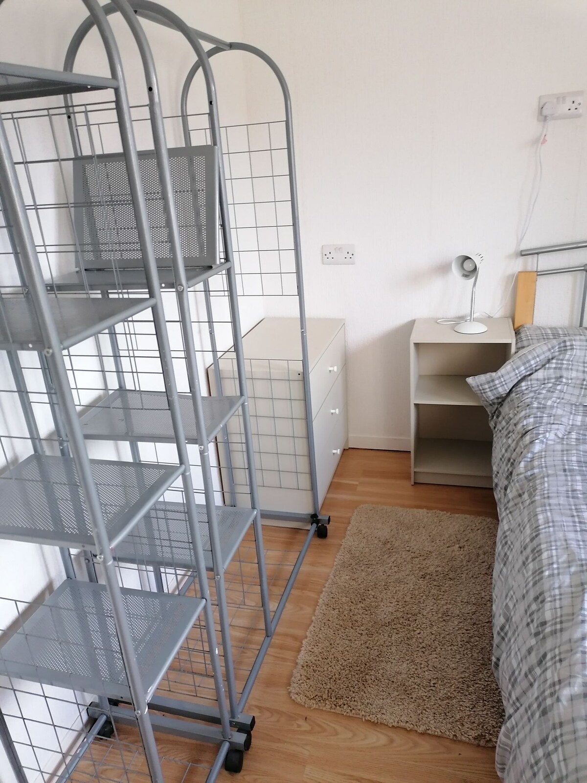 A compact bedroom features a patterned bedspread on a spacious bed, complemented by bedside furniture. A metal shelving unit and a small cabinet provide storage options, while a desk lamp on a nightstand offers additional lighting. A beige area rug is placed on the wooden floor.