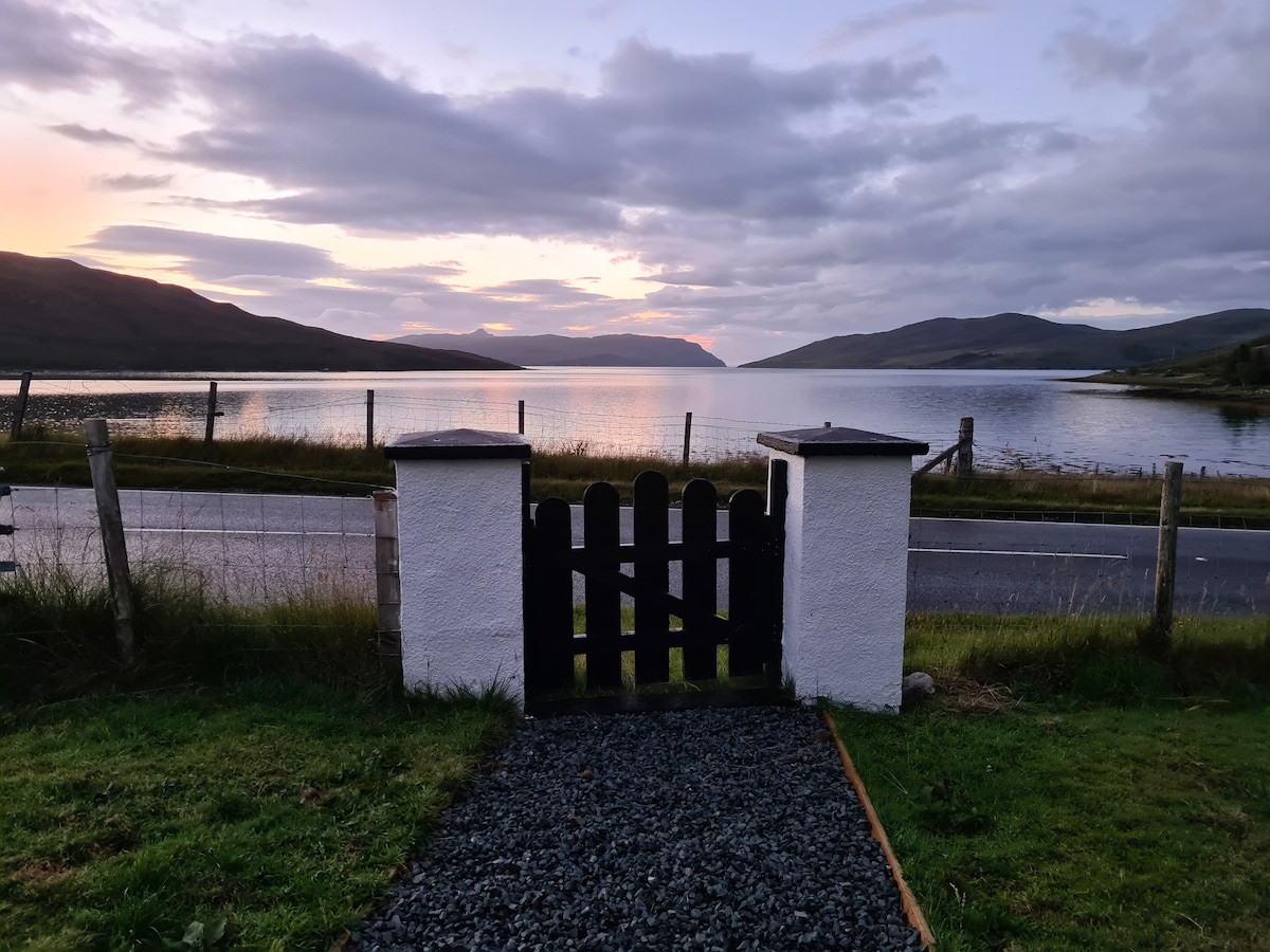 A gate opens from a gravel path towards a stunning view of Loch Ainort. The calm waters reflect the evening sky, while hills rise softly in the background. The surrounding grass is neatly maintained, and a fence lines the property along the roadside.