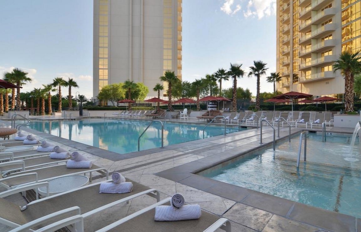 Two pool areas are featured, surrounded by palm trees and lounge chairs. Umbrellas provide shade to guests. Water features can be seen in the foreground, while tall buildings rise in the background under a clear sky.