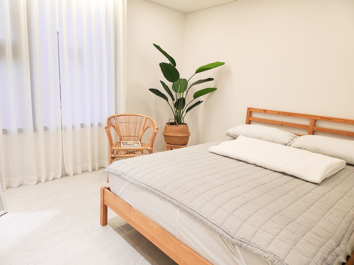 A well-lit bedroom features a spacious bed with soft linens, complemented by a minimalist design. A large indoor plant adds a touch of greenery, while a rattan chair sits comfortably in the corner. Sheer curtains allow natural light to filter softly into the room.