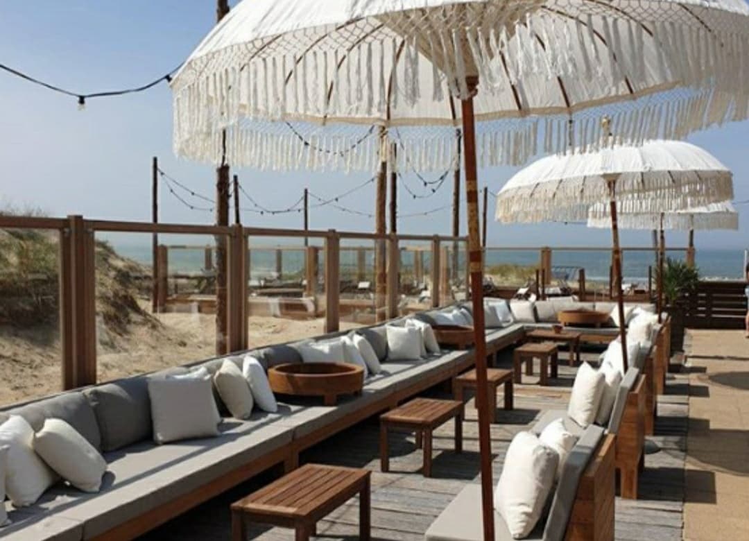 An outdoor lounge area features a series of wooden benches with soft cushions, complemented by small wooden tables. Large umbrellas provide shade, while the ocean is visible in the background. Warm lighting from string lights enhances the relaxing environment.