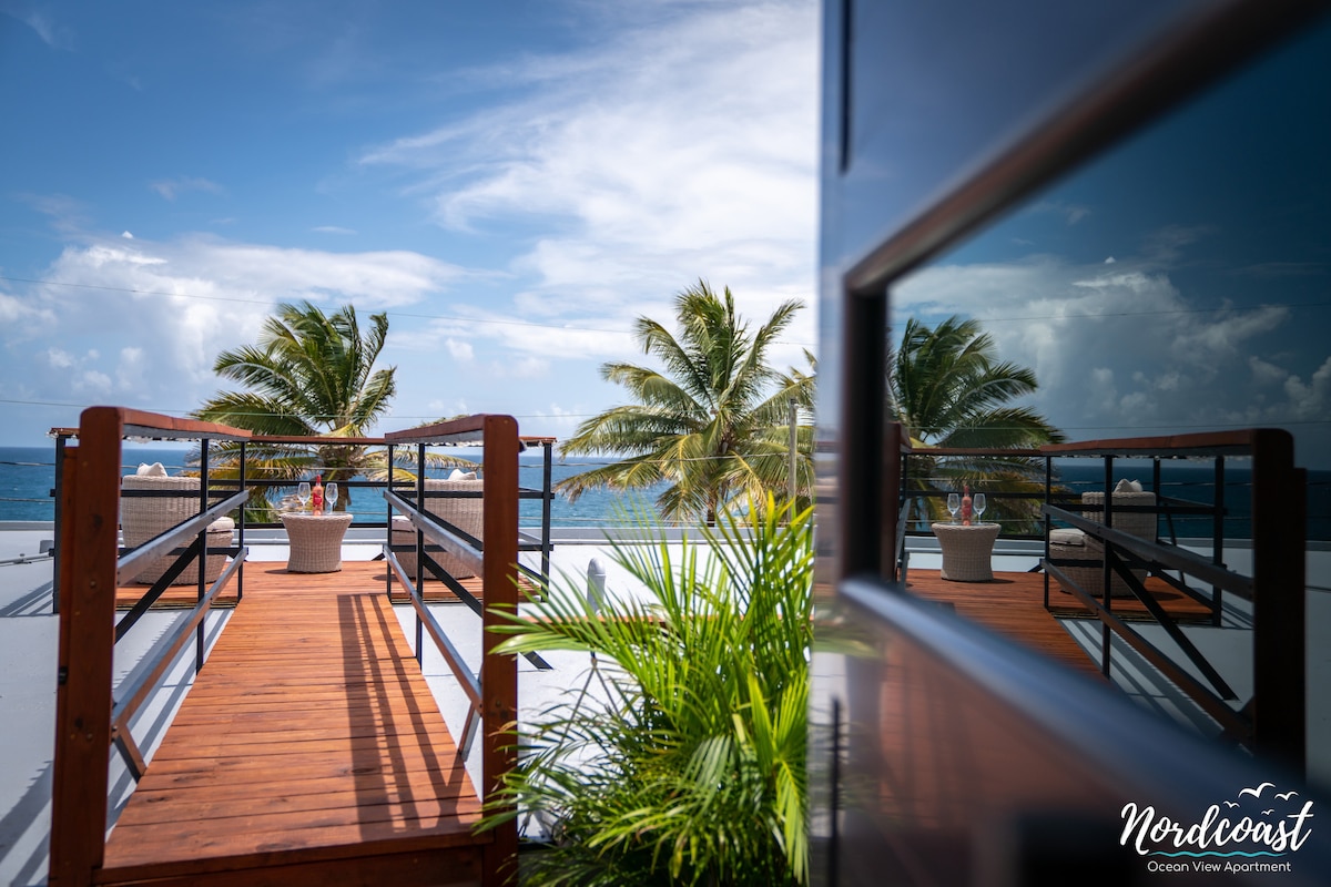 A wooden deck extends towards the ocean, framed by palm trees. Reflections can be seen in a nearby glass surface, highlighting the tranquil atmosphere. Clear blue skies complement the serene waters, creating a welcoming outdoor space for relaxation.