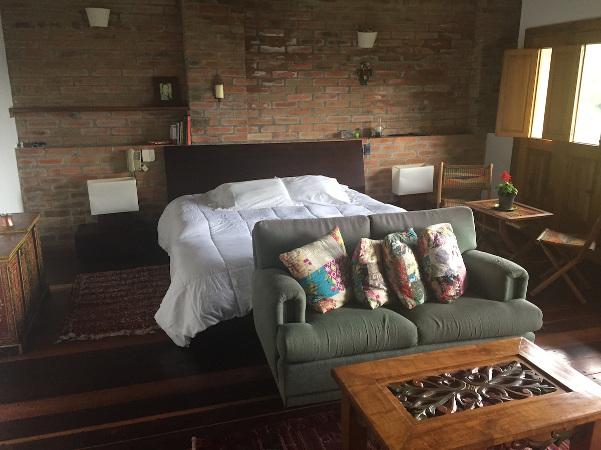 A spacious bedroom features a large bed with soft linens, complemented by a cozy sofa adorned with decorative pillows. Wooden furniture pieces add warmth to the room, while natural light filters through the windows, enhancing the overall ambiance.