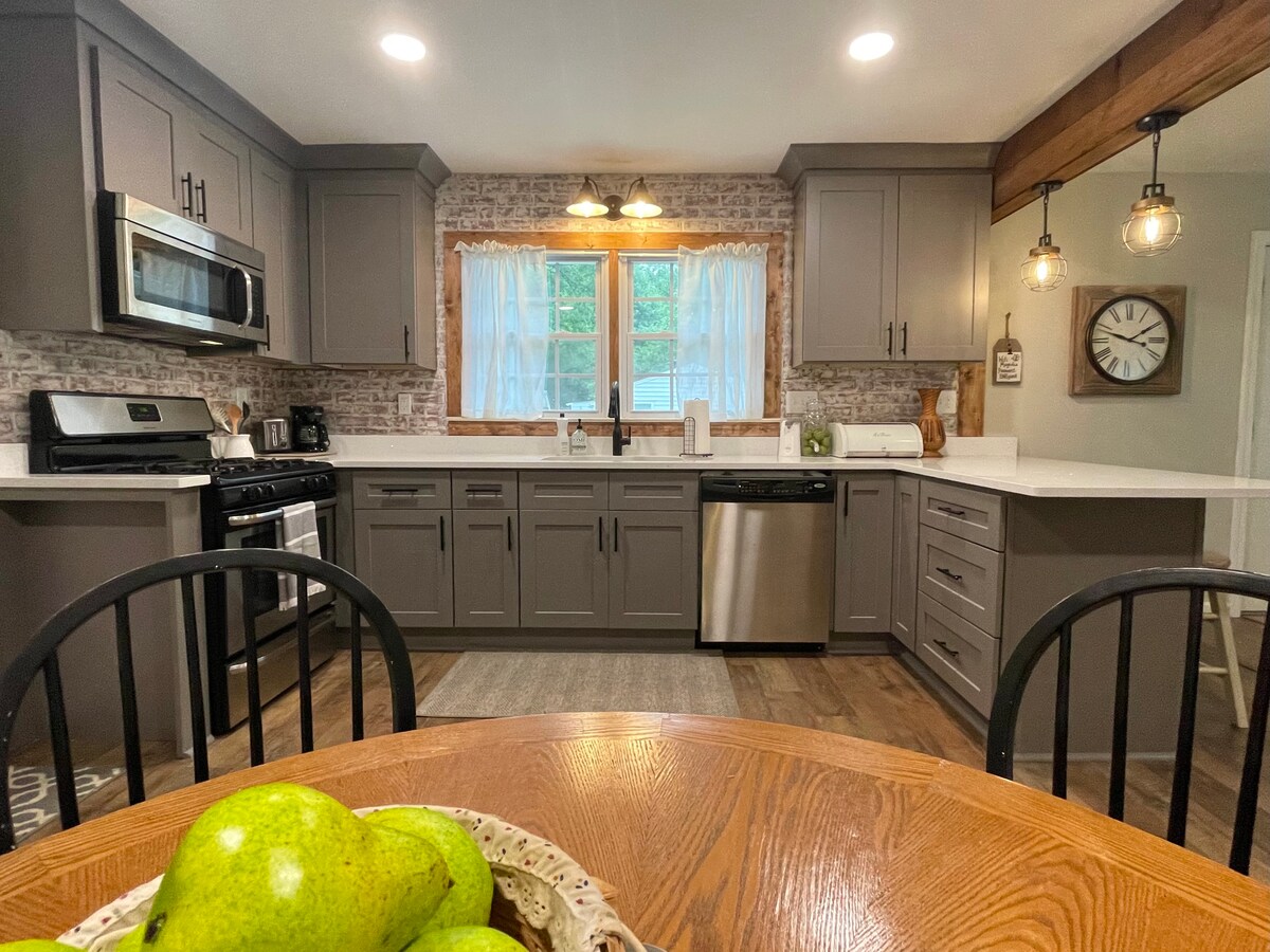 The kitchen features modern gray cabinetry and a white countertop. Stainless steel appliances include a microwave and dishwasher. A window above the sink allows natural light, and decorative pendant lights hang from a wooden beam. A wooden dining table is set with a bowl of green apples.